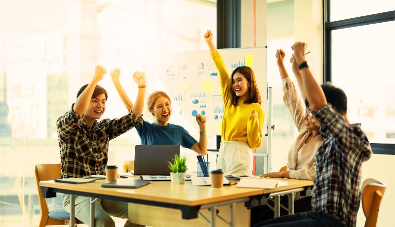 A group of five people sitting around a desk in a brightly lit office, celebrating with raised arms, smiling, and appearing excited, with charts and a whiteboard in the background.