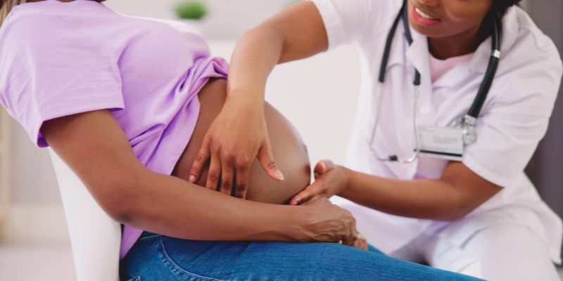 A pregnant woman wearing a purple top and blue jeans sits while a healthcare provider, dressed in white with a stethoscope, gently examines her baby bump