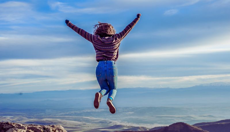 A person jumps in the air with arms raised while wearing a striped sweater and blue jeans..