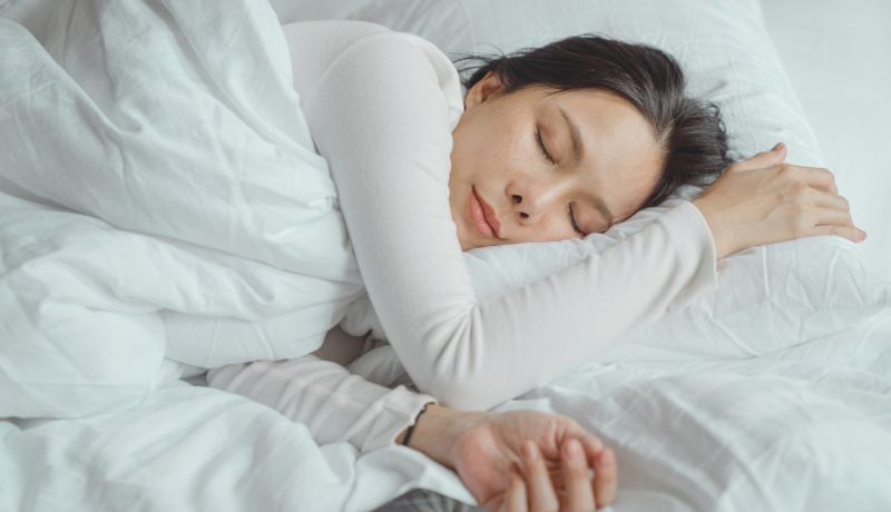 A woman wearing long sleeved white shirt covered with a white blanket lying in bed sleeping.