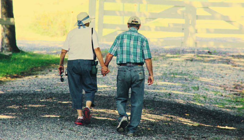 Two people walking hand in hand on a gravel path in an outdoor setting with trees and a white fence in the background.